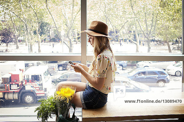 Seitenansicht einer Frau  die telefoniert  während sie an Blumen und Glasfenster sitzt