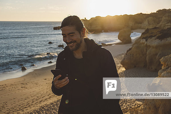 Lächelnder Mann benutzt Mobiltelefon  während er bei Sonnenuntergang am Strand steht