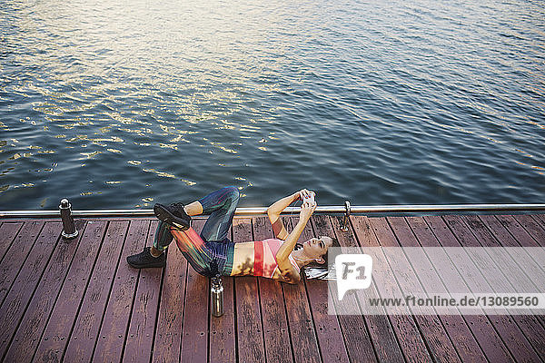 High angle view of woman lying and using smart phone on wooden walkway by river
