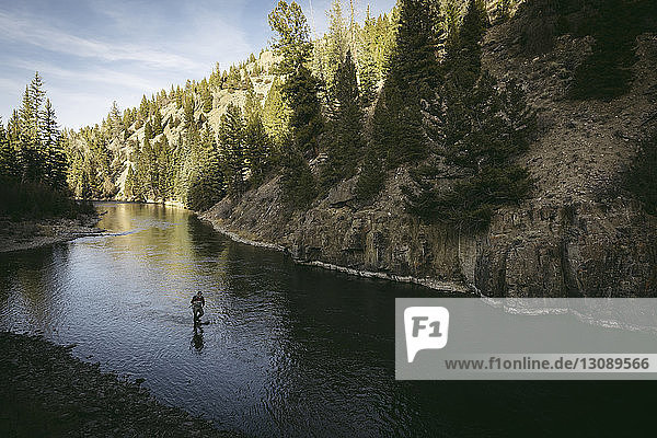 Hochwinkelaufnahme eines Mannes  der im Fluss inmitten eines Berges im Wald fischt