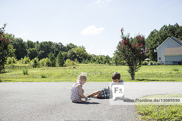 Siblings writing with chalk on road against sky during sunny day