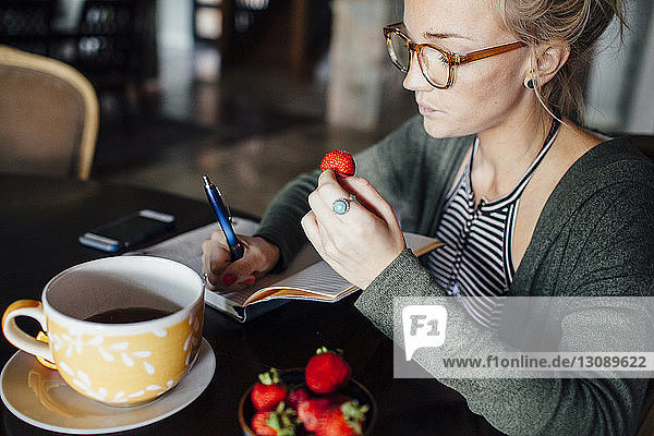 Woman with strawberry writing diary while sitting at table