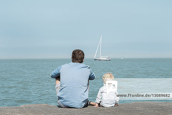 Rückansicht eines Vaters mit Sohn am Strand sitzend vor blauem Himmel am sonnigen Tag