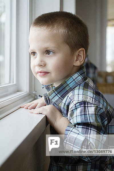 Lächelnder Junge schaut durchs Fenster  während er zu Hause steht