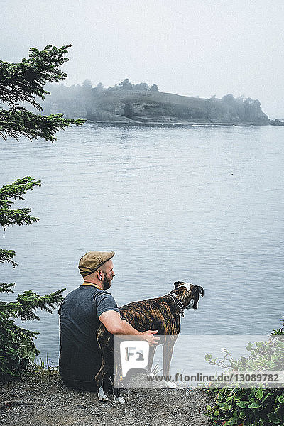 Rückansicht eines am See sitzenden Mannes mit Hund