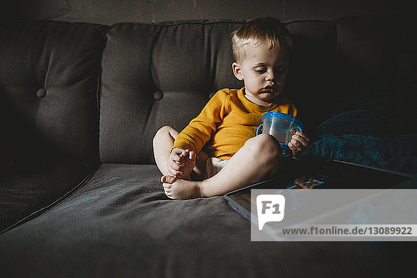 Cute baby boy with mug sitting on sofa at home