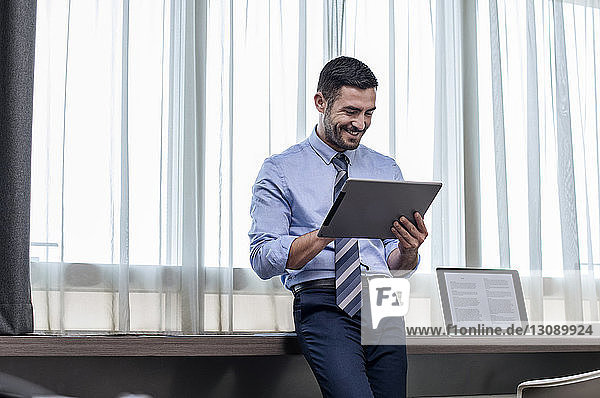 Happy businessman using tablet computer while standing by window in hotel room