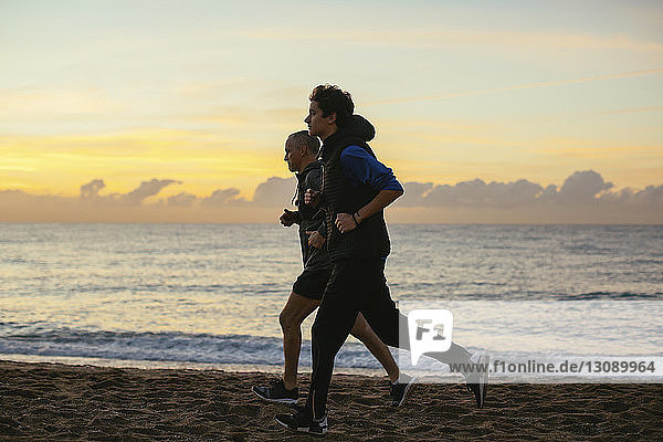 Determined father and son jogging at beach against cloudy sky during sunset