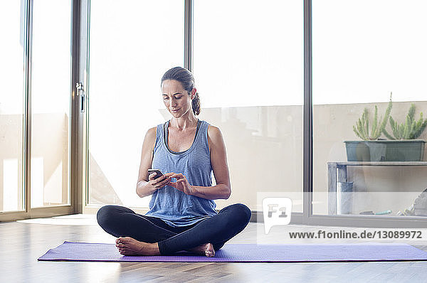 Frau benutzt Smartphone  während sie zu Hause auf der Gymnastikmatte sitzt