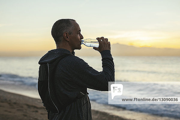 Seitenansicht eines Mannes  der bei Sonnenuntergang Wasser trinkt  während er am Strand steht