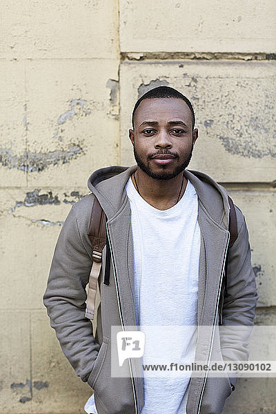 Portrait of confident young man standing with hands in pockets against wall