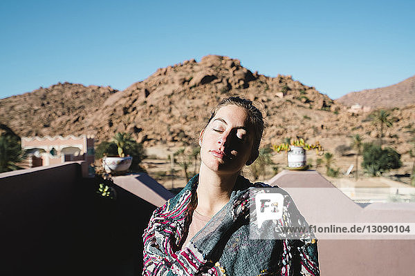Frau mit geschlossenen Augen steht an sonnigen Tagen auf der Terrasse vor den Bergen
