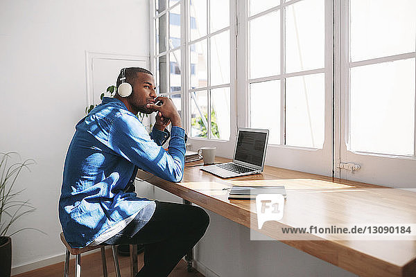 Seitenansicht eines nachdenklichen Mannes  der Musik hört  während er mit einem Laptop-Computer an einem Holztisch am Fenster sitzt