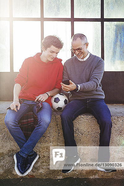 Happy father showing smart phone to son while sitting on retaining wall