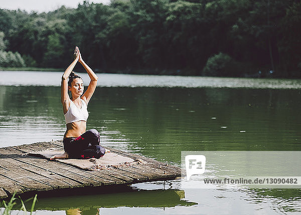 Frau mit erhobenen Armen meditiert  während sie auf einem Steg über einem See im Wald sitzt