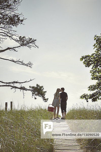 Rear view of couple kissing while standing on boardwalk amidst field against sky