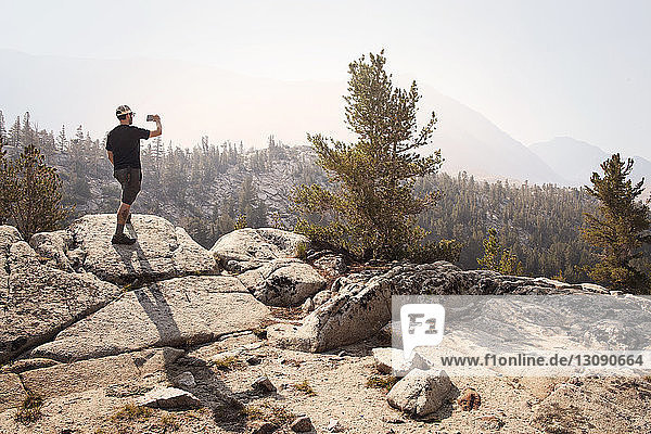 Rückansicht eines Mannes  der auf einem Fels steht und an einem sonnigen Tag gegen Berge fotografiert