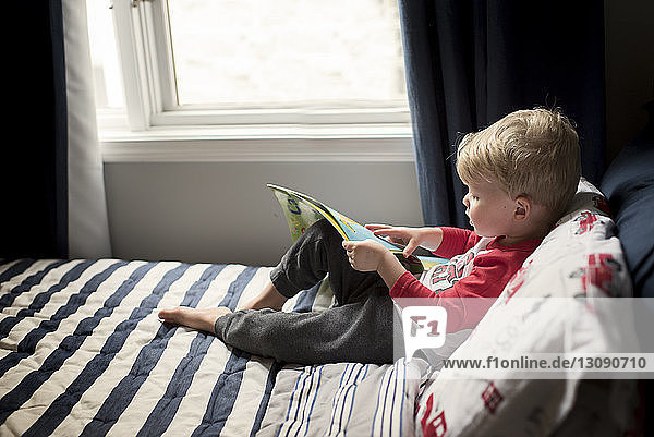 Hochwinkelansicht eines Jungen  der ein Bilderbuch hält  während er zu Hause auf dem Bett sitzt
