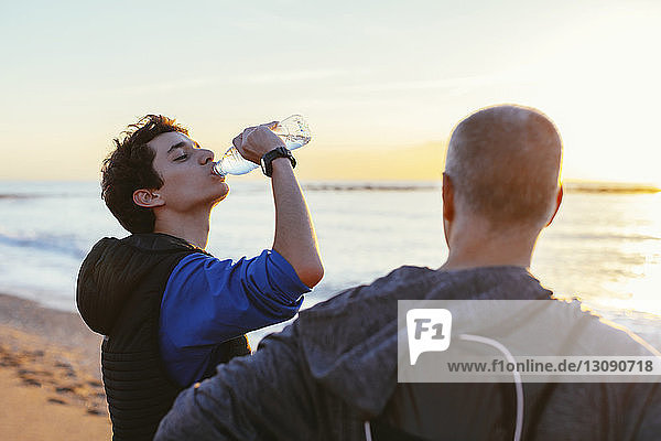 Son drinking water while standing by father at beach during sunset