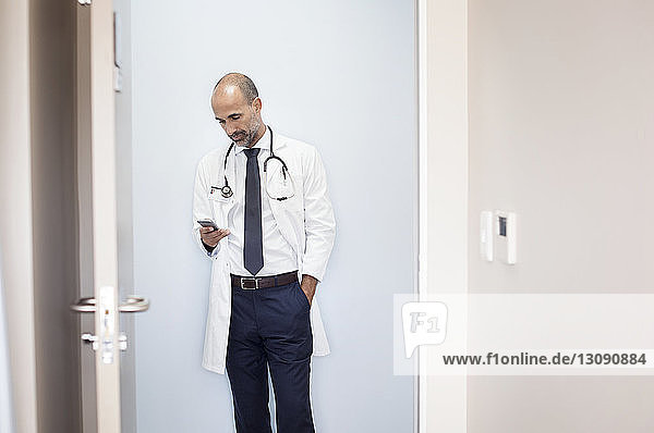 Doctor using smart phone while standing against wall at hospital seen through door