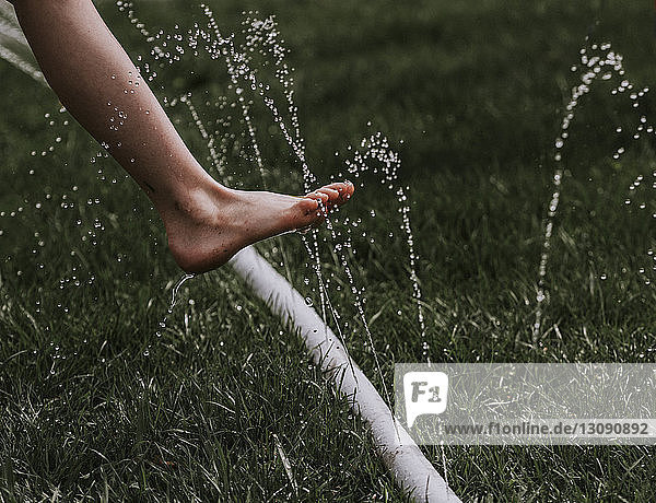 Low section of boy over water spraying garden hose on grassy field at backyard