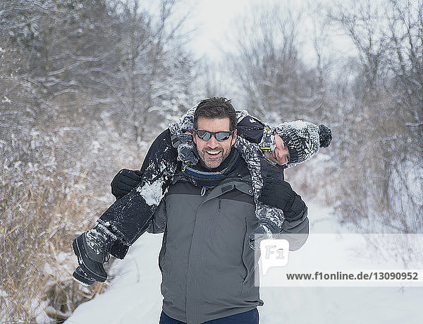 Playful father carrying son on shoulders at snowy field against bare trees