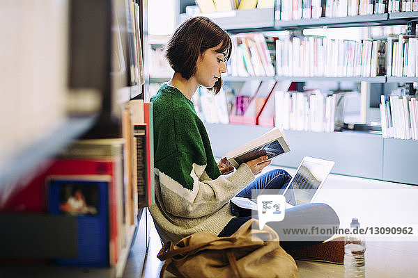 Seitenansicht einer Frau mit Laptop-Computer  die in der Bibliothek auf dem Boden sitzend ein Buch liest