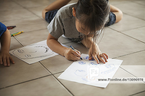 Girl drawing on paper while lying besides brother on floor at home