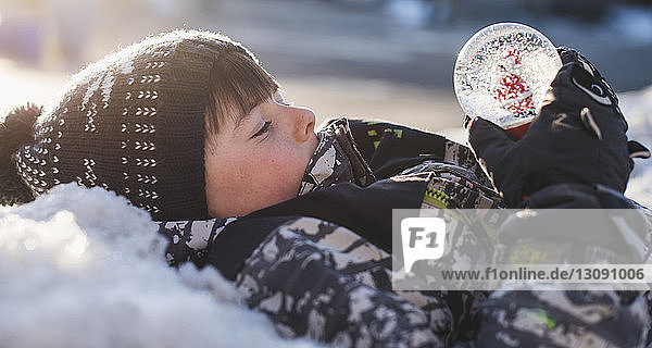 Side view of boy looking at snow globe while lying outdoors during winter