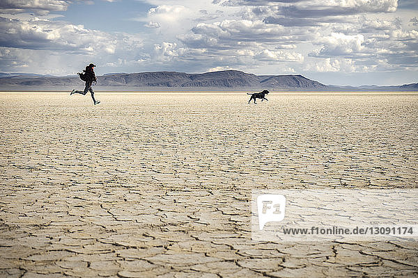 Woman and dog running on dry field against cloudy sky at Alvord Desert