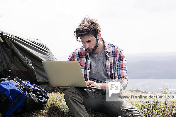 Man using laptop at campsite on hill against clear sky