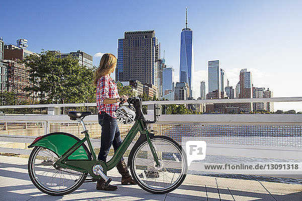 Frau mit Fahrrad geht auf Brücke und schaut Fluss gegen Stadt