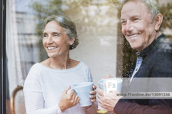 Happy senior couple holding coffee mugs while looking through window at home