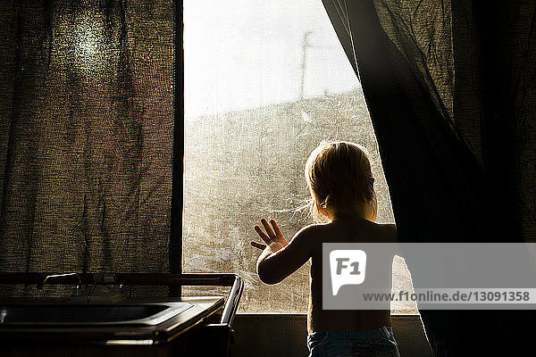 Rear view of boy looking through window while standing at home