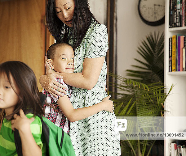Mutter umarmt Sohn  während im Vordergrund ein Mädchen mit Rucksack steht