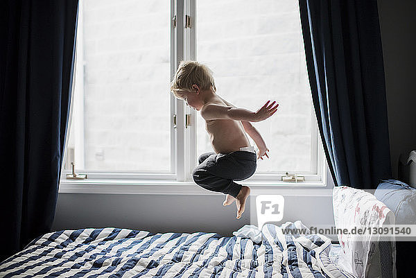 Shirtless boy jumping on bed by window at home