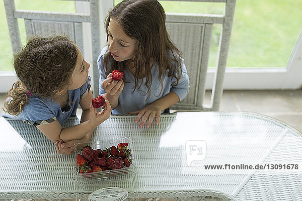 High angle view of sisters looking face to face while eating strawberries at table