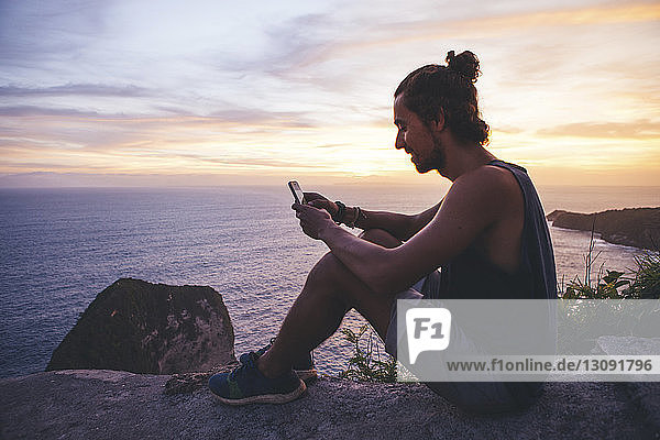 Seitenansicht eines Mannes  der ein Mobiltelefon benutzt  während er bei Sonnenuntergang auf einem Berg über dem Meer gegen den Himmel sitzt