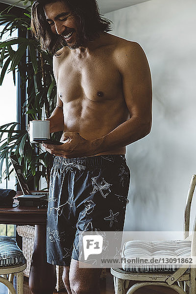 Mann ohne Hemd hält Becher  während er zu Hause ein Mobiltelefon benutzt