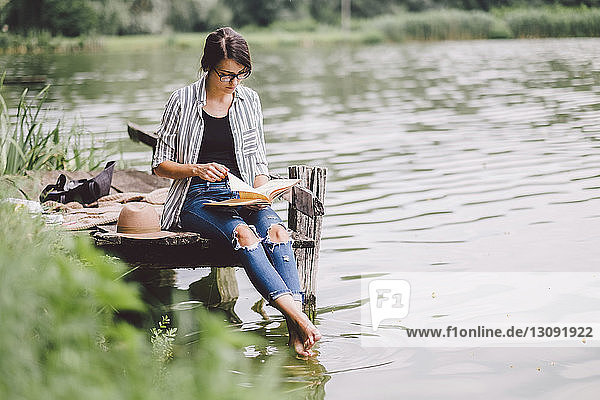 Frau liest Buch  während sie auf dem Pier über dem See im Wald sitzt