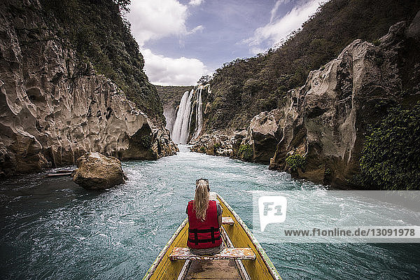 Rear view of woman sitting in boat on river by mountains against waterfall