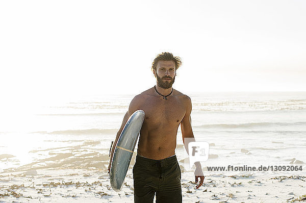 Portrait of shirtless man carrying surfboard while walking at beach against clear sky
