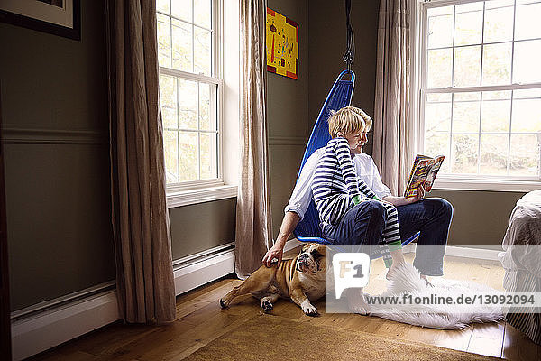 Father reading book to son and stroking dog while sitting on swing at home