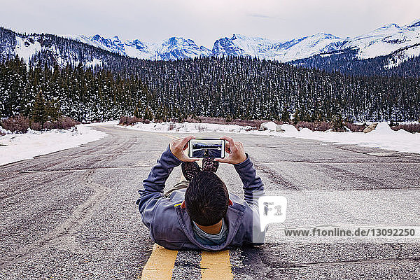 Mann liegt auf der Straße und fotografiert mit Smartphone vor schneebedeckten Bergen