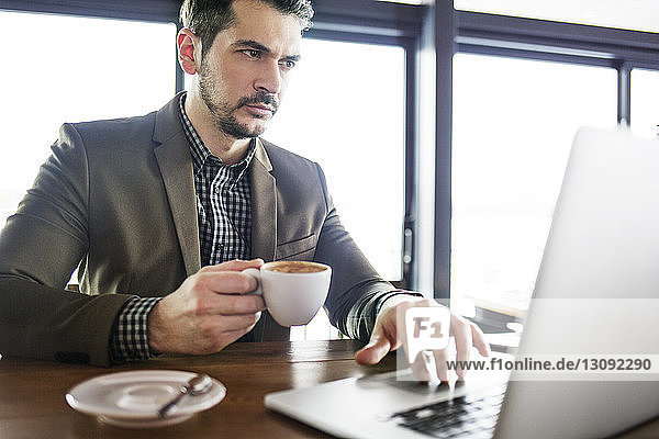 Concentrated businessman holding coffee cup while working on laptop in cafe