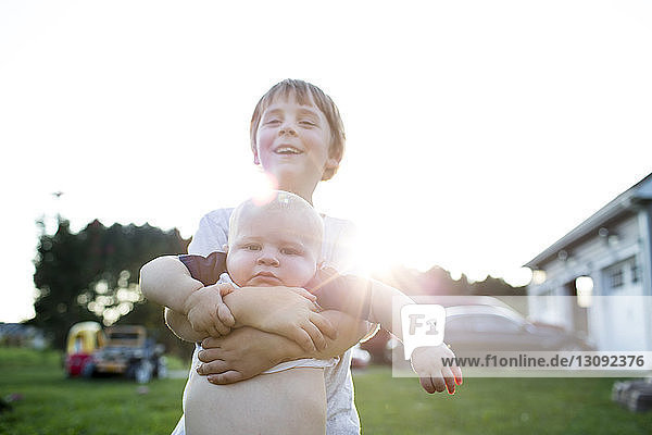 Smiling boy carrying brother while standing at backyard against clear sky