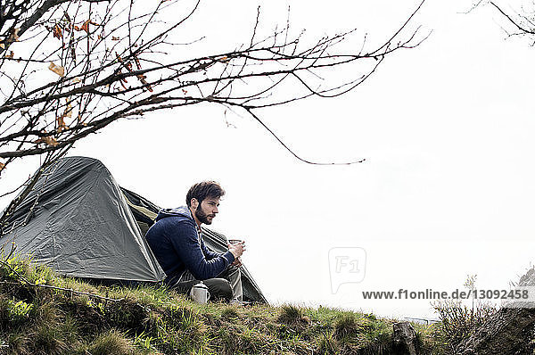 Nachdenklicher Mann sitzt vor dem Zelt auf einem Hügel vor klarem Himmel