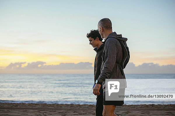 Side view of father and son walking at beach against sky