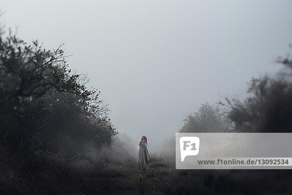 Girl standing on field against clear sky while being wrapped in blanket during foggy weather