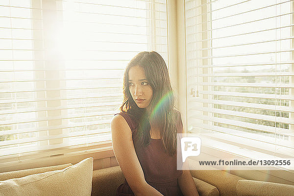Nachdenkliche Frau sitzt an sonnigen Tagen auf dem Sofa am Fenster